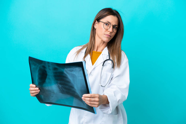 Mujer de mediana edad aislada sobre fondo azul con una bata de médico y sosteniendo una gammagrafía ósea - Foto, imagen