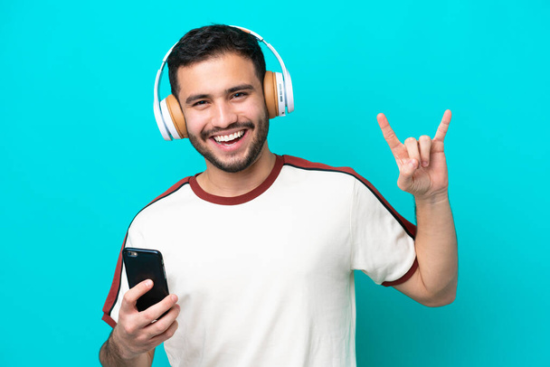Joven brasileño aislado sobre fondo azul escuchando música con un móvil haciendo gestos de rock - Foto, imagen
