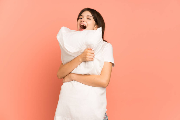 Молода українська дівчина ізольована на рожевому тлі в піжамі і тримає подушку з радісним виразом обличчя. - Фото, зображення