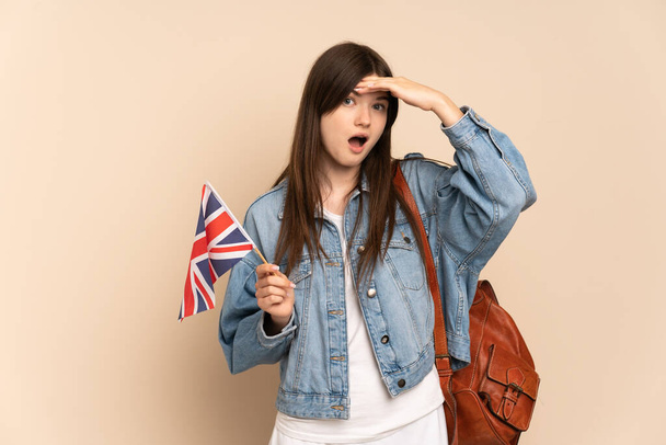 Ukraynalı genç bir kız bej arka planda izole edilmiş bir Birleşik Krallık bayrağı tutuyor. Yan tarafa bakarken sürpriz jest yapıyor. - Fotoğraf, Görsel