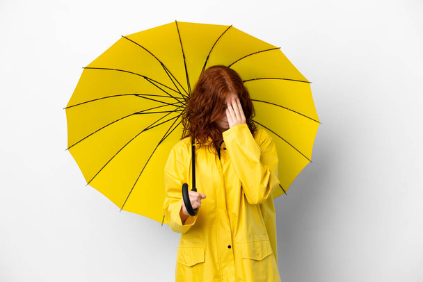 Genç kızıl saçlı kız yağmurluğa ve şemsiyeye dayanıklı beyaz arka planda, yorgun ve hasta bir ifadeyle izole edilmiş. - Fotoğraf, Görsel