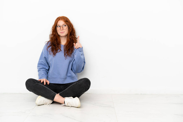 Tini vörös hajú lány ül a padlón elszigetelt fehér háttér gondolkodás egy ötlet rámutatva az ujját fel - Fotó, kép