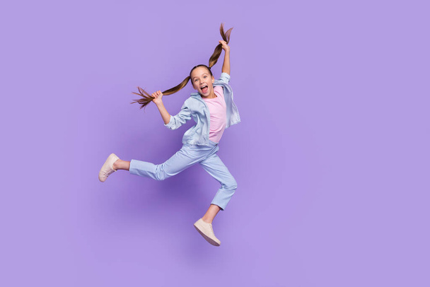 Foto de muito funky menina da escola usar camisa jeans saltando alta segurando caudas espaço vazio isolado cor roxa fundo - Foto, Imagem