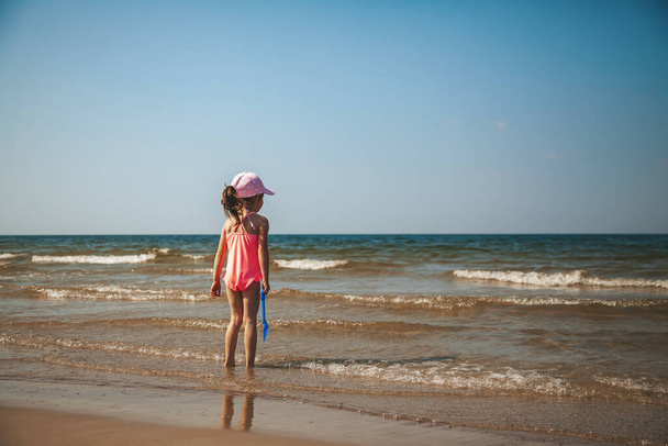Cute little girl in a pink swimsuit watching the sea standing on the beach - Foto, Imagen