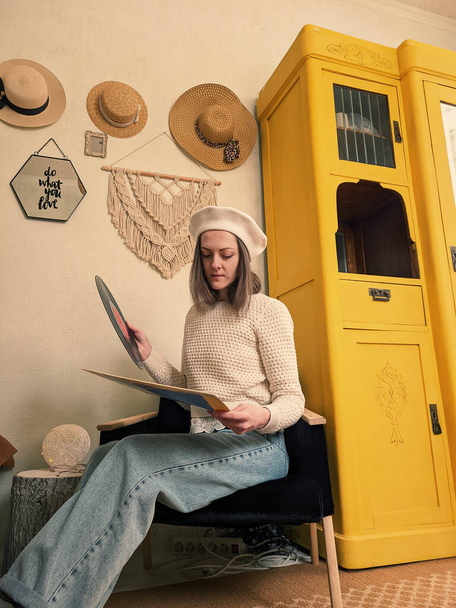 Kharkiv, Ucrania. 01.22.2022. Mujer joven en boina se sienta en un sillón en casa acogedora, en la habitación con registro de música en las manos. Armario amarillo vintage. Interior del hogar - Foto, imagen
