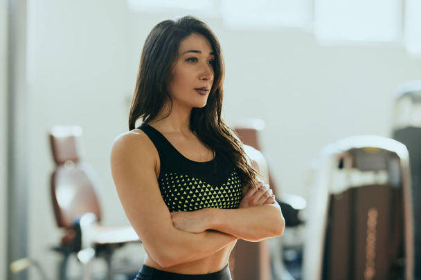 Belle sportive brune regardant la caméra avec les bras croisés dans une salle de gym. - Photo, image