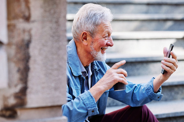 Un homme âgé joyeux fait un appel vidéo avec sa famille sur son téléphone alors qu'il est assis dans les escaliers à l'extérieur. - Photo, image