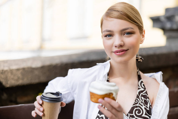 young blonde woman with cupcake and paper cup smiling at camera outdoors - Photo, Image