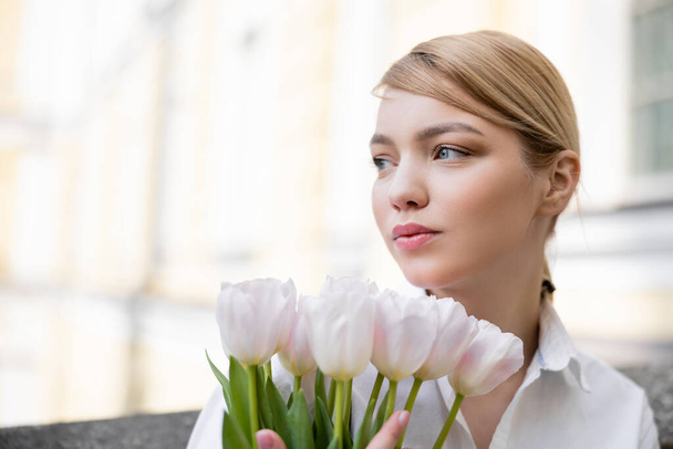 jovem e encantadora mulher olhando para longe perto de tulipas brancas ao ar livre - Foto, Imagem