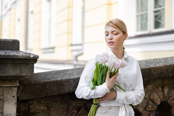 vrouw in wit shirt staan met boeket van tulpen in de buurt van stenen hek - Foto, afbeelding