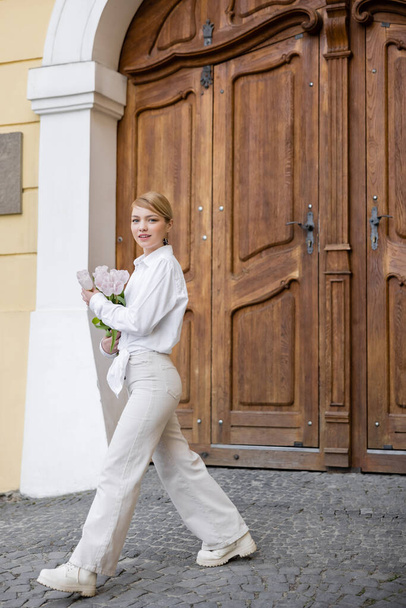visão de comprimento total da mulher elegante com tulipas andando perto do edifício com portas de madeira - Foto, Imagem