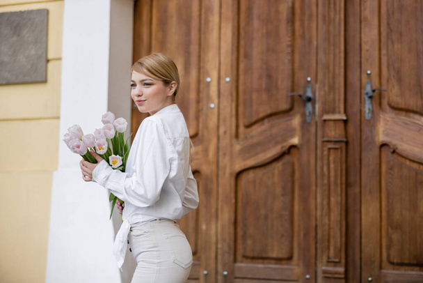 stylish and smiling woman standing with bouquet of flowers near blurred wooden doors - Fotografie, Obrázek