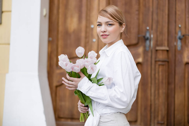 довольная женщина в белой рубашке с букетом тюльпанов на городской улице - Фото, изображение