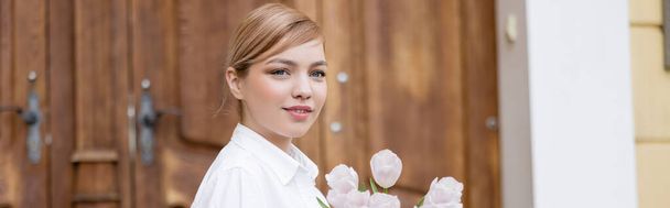 blonde young woman smiling near fresh tulips outdoors, banner - 写真・画像