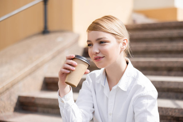 pleased woman in white shirt holding coffee to go outdoors - Фото, изображение