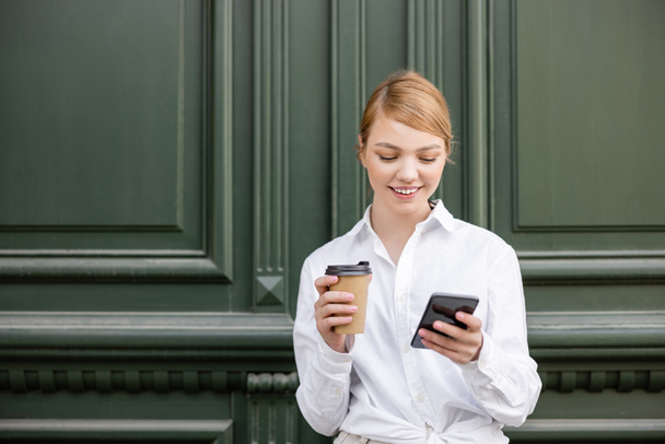 cheerful young woman with takeaway drink looking at mobile phone near grey wall - Photo, image