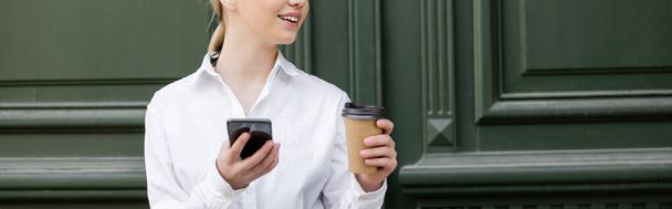 cropped view of woman with paper cup and smartphone smiling near grey wall, banner - Photo, Image