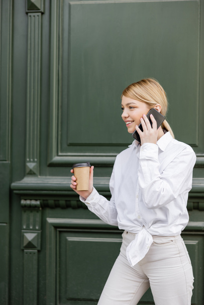 szczęśliwa kobieta dzwoni na telefon komórkowy, stojąc z papierową filiżanką w pobliżu szarej ściany - Zdjęcie, obraz
