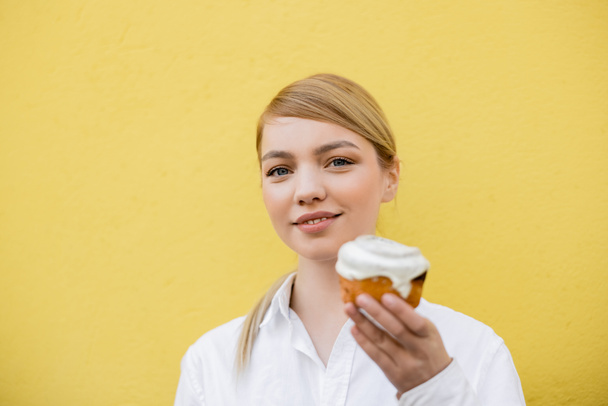 glückliche junge Frau mit Cupcake blickt in die Kamera isoliert auf gelb - Foto, Bild
