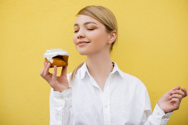 pleased woman in white shirt holding cupcake isolated on yellow - Foto, Bild