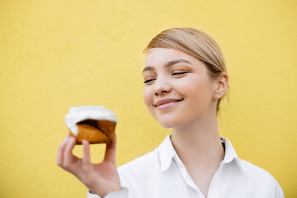 cheerful blonde woman holding tasty cupcake isolated on yellow - Photo, image