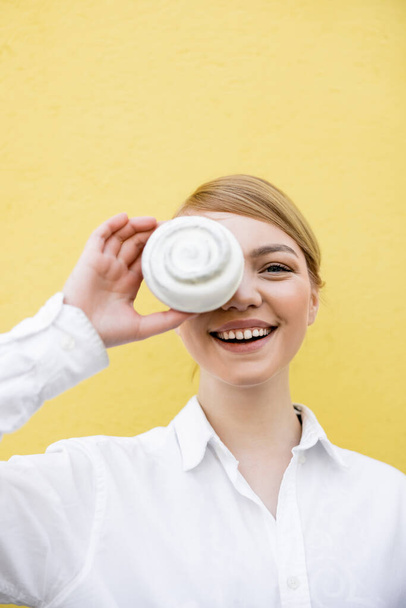 cheerful woman covering eye with cupcake while smiling at camera isolated on yellow - Foto, Imagem
