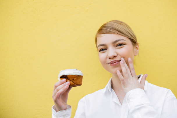 glückliche Frau hält köstliche Cupcake und wischt Lippen isoliert auf gelb - Foto, Bild