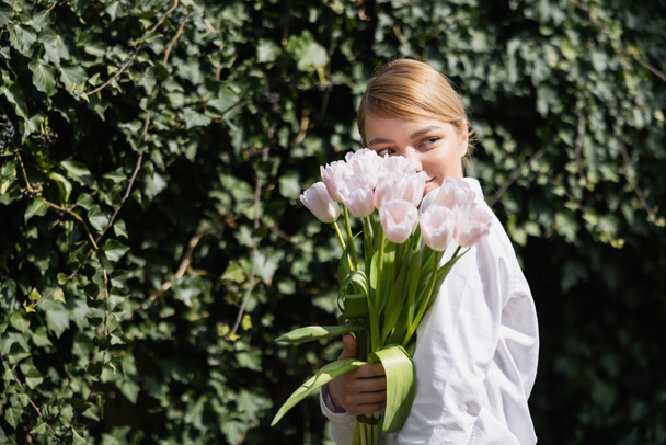 heureuse jeune femme obscurcissant visage avec des tulipes blanches près de lierre vert - Photo, image