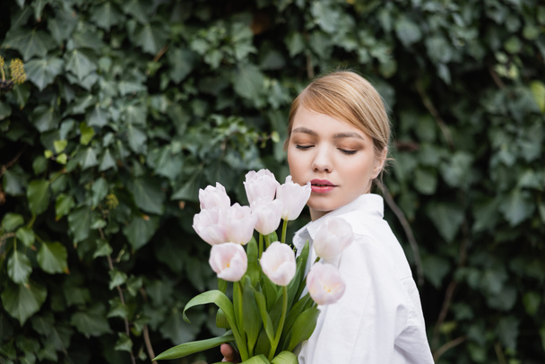 charming woman with bouquet of white tulips standing near green ivy - 写真・画像