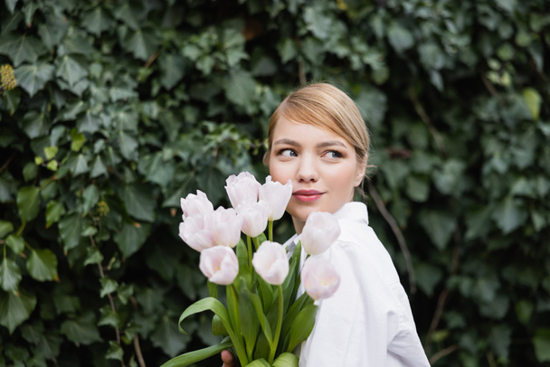 jeune femme blonde avec des tulipes blanches souriant près de lierre vert à l'extérieur - Photo, image