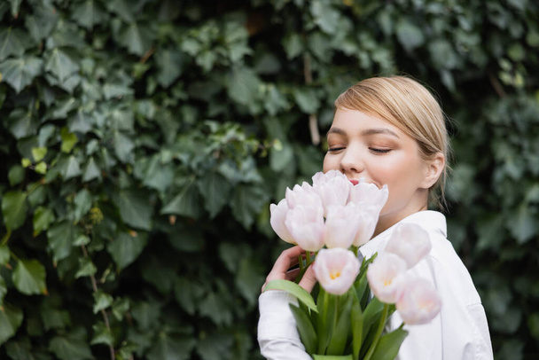счастливая молодая женщина скрывает лицо с белыми тюльпанами рядом с зеленым плющом - Фото, изображение