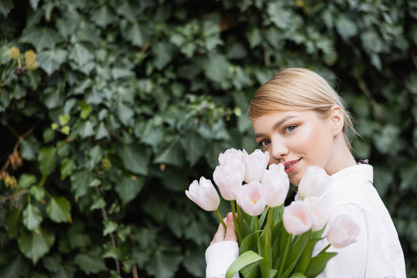 charmante vrouw met witte tulpen glimlachen op camera in de buurt wazig klimop - Foto, afbeelding