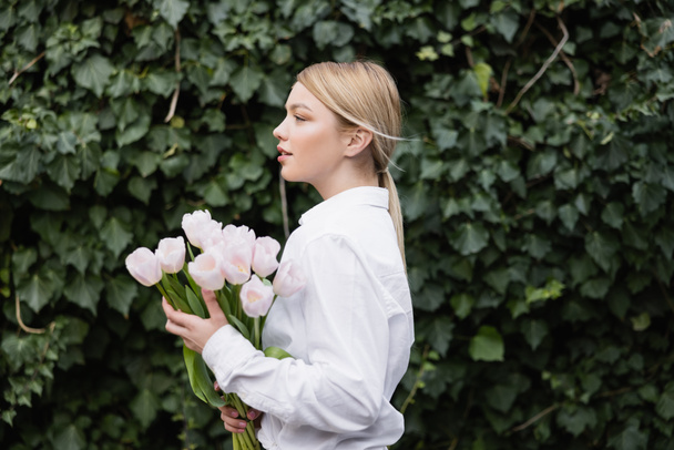 Seitenansicht einer blonden Frau mit weißen Tulpen, die neben grünem Efeu steht - Foto, Bild