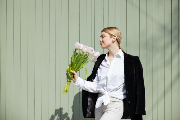 pleased woman in white shirt and black blazer standing with tulips near grey wall - Foto, Bild