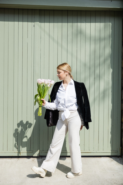 full length view of trendy woman with bouquet of white tulips near grey wall outdoors - Фото, изображение