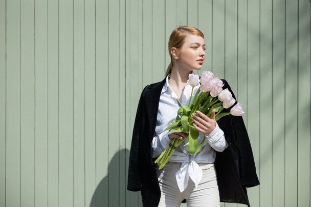 молода жінка в білій сорочці і чистий блейзер стояв з тюльпаном біля сірої стіни. - Фото, зображення
