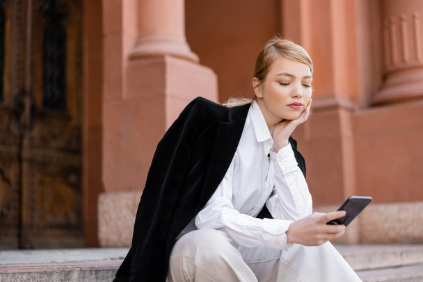 mujer joven con camisa blanca y chaqueta negra usando smartphone en la calle urbana - Foto, Imagen