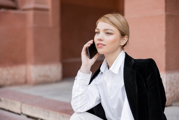 mujer bastante joven llamando en el teléfono inteligente mientras está sentado en la calle de la ciudad - Foto, imagen