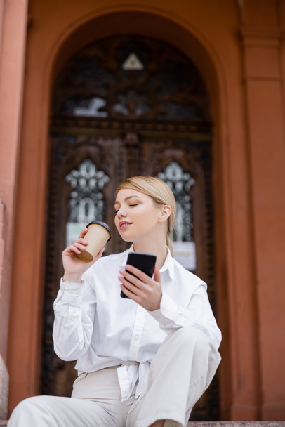 blonde woman with smartphone drinking coffee near blurred building - Фото, изображение