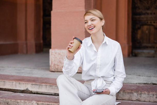 cheerful young woman sitting on stair with mobile phone and coffee to go - Φωτογραφία, εικόνα
