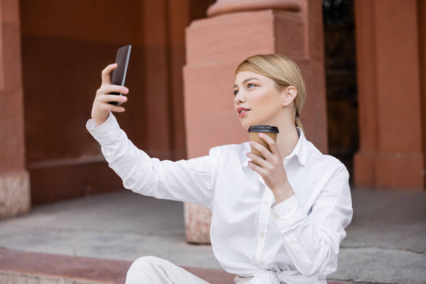 κομψή γυναίκα με καφέ για να πάει τη λήψη selfie στο κινητό τηλέφωνο σε εξωτερικούς χώρους - Φωτογραφία, εικόνα