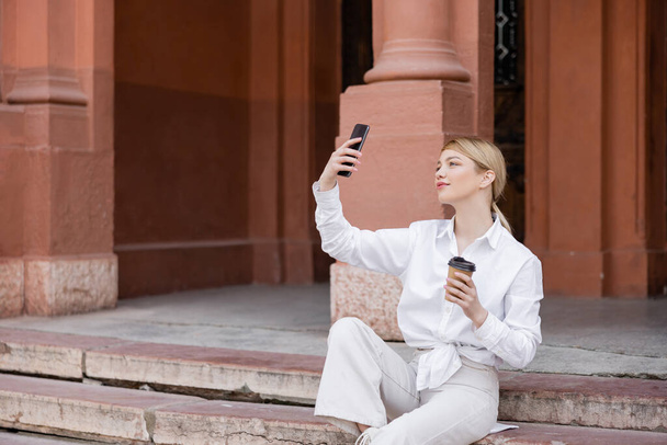 όμορφη γυναίκα με καφέ για να πάει κάθεται στις σκάλες και να παίρνει selfie στο κινητό - Φωτογραφία, εικόνα