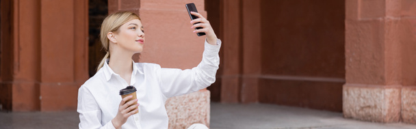 charming woman with takeaway drink taking selfie on smartphone outdoors, banner - 写真・画像