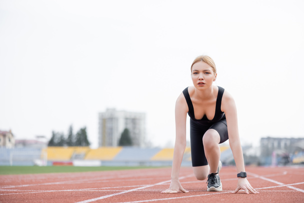 young sportswoman looking at camera while standing in low start position on stadium - Photo, Image