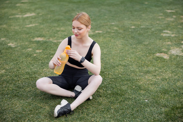 Genç sporcu kadın stadyumda oturuyor ve spor şişesini açıyor. - Fotoğraf, Görsel