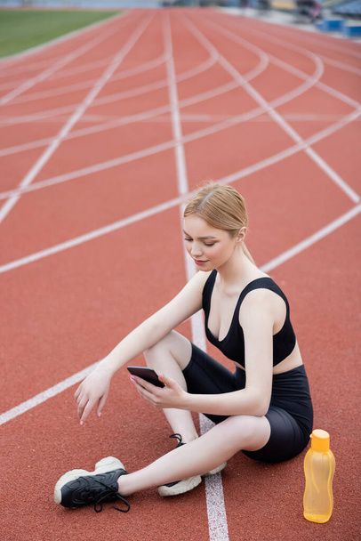 jovem mulher esportiva sentado com pernas cruzadas e usando celular - Foto, Imagem