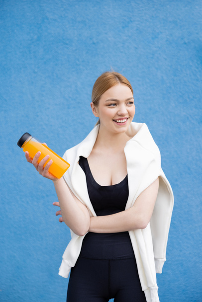 joyful sportswoman looking away while standing with orange juice on blue textured background - Foto, Imagem