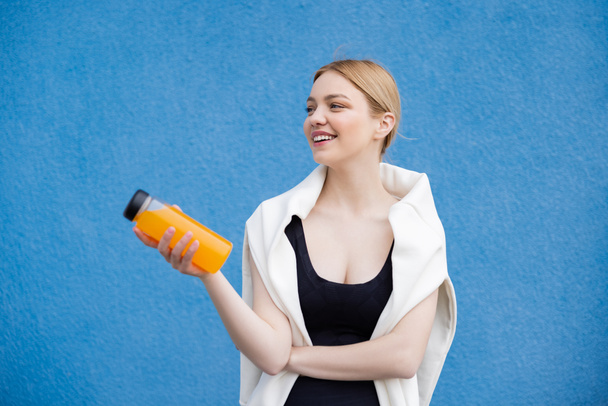 vrolijke vrouw in sportkleding met oranje sap op blauwe textuur achtergrond - Foto, afbeelding