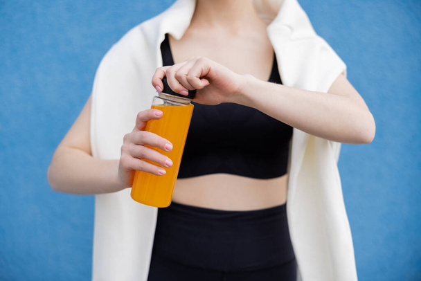 cropped view of blurred woman in sportswear opening bottle with orange juice on blue background - Foto, afbeelding