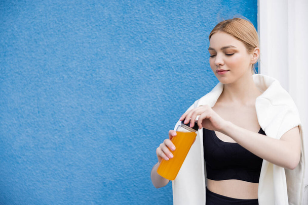 młoda i sportowa kobieta otwiera butelkę soku pomarańczowego w pobliżu niebieskiej teksturowanej ściany - Zdjęcie, obraz